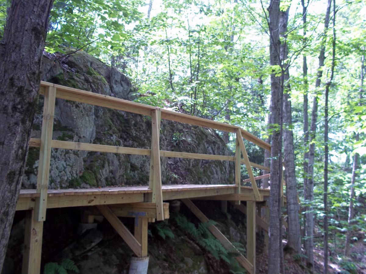 A wooden boardwalk with a railing leads through a wooded area, featuring a large rock in the background and surrounded by lush green foliage. Sunlight filters through the trees, creating a serene and inviting natural setting. Noquemanon Trails Network: South Marquette Trails mountain bike trail.