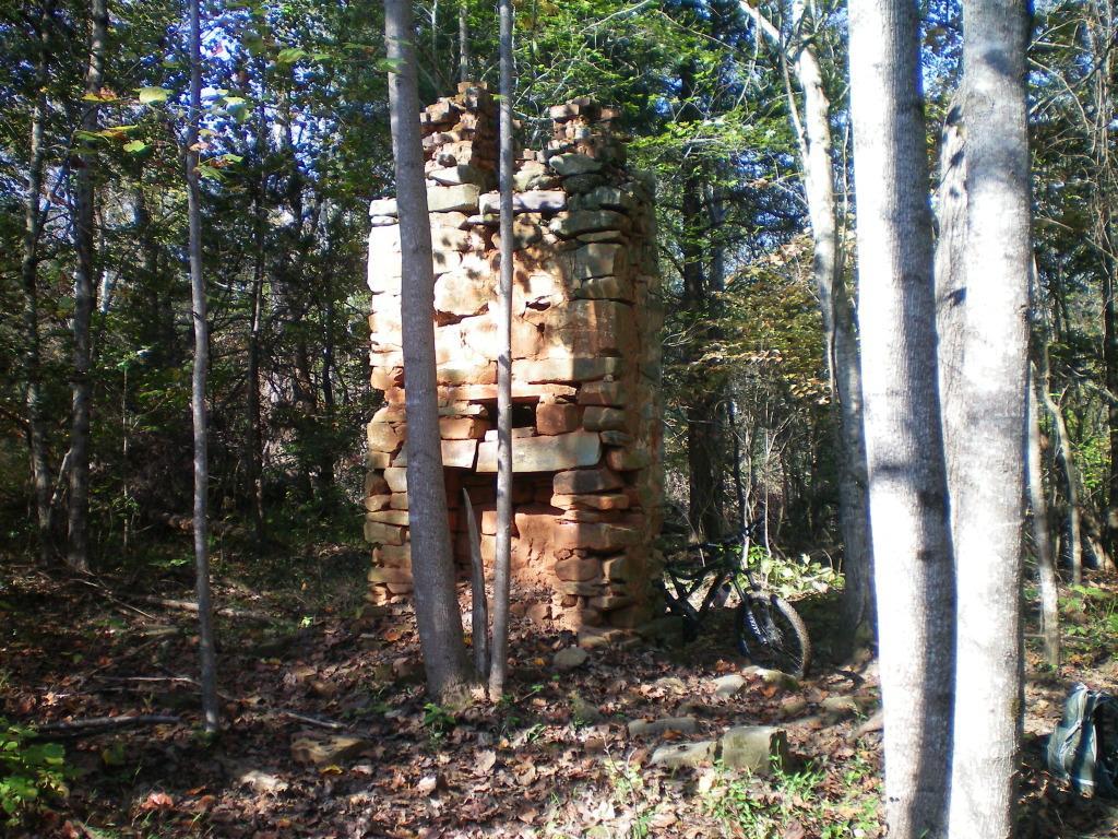 Alt text: A weathered stone chimney structure surrounded by trees in a forested area, with fallen leaves on the ground and a bicycle partially visible in the foreground. Walnut Creek mountain bike trail.