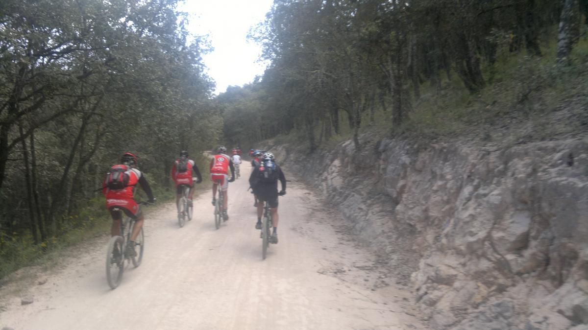 A group of mountain bikers riding along a dirt trail surrounded by trees in a natural setting. The riders are wearing red and black cycling gear and are positioned in a line, moving away from the camera. The trail is flanked by rocky terrain and greenery. Tierra Blanca - Derramadero mountain bike trail.