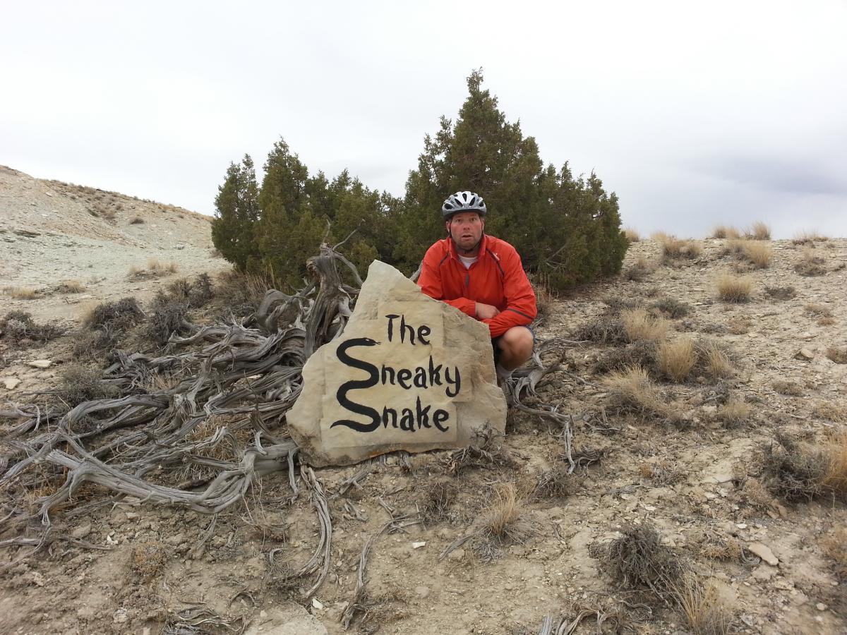 A person wearing a helmet and an orange jacket poses next to a large stone sign that reads "The Sneaky Snake." The background features a rugged, dry landscape with sparse vegetation and a cloudy sky. Tnt mountain bike trail.
