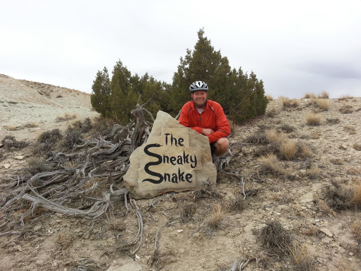 A man in a red jacket and bike helmet smiles beside a large rock with the inscription "The Sneaky Snake" in a desert landscape. Scrub trees and dry grass are visible in the background under a cloudy sky. Tnt mountain bike trail.
