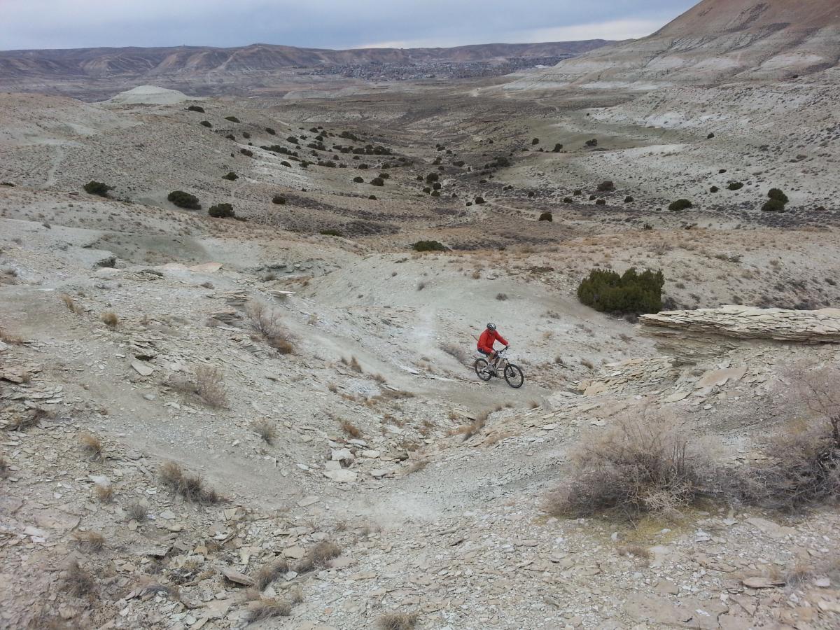 A person riding a mountain bike down a rocky, sloped trail in a vast, barren landscape. The terrain features dry ground, small shrubs, and distant hills under a cloudy sky. Tnt mountain bike trail.