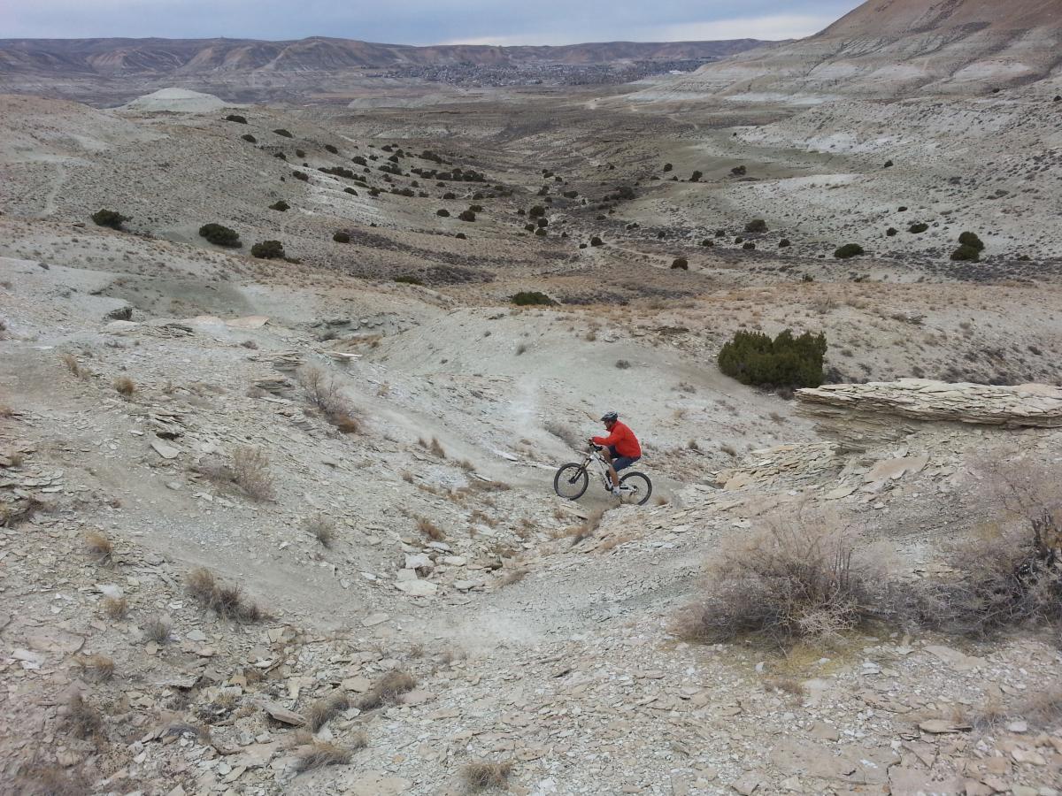 A mountain biker in a bright red jacket navigates a rocky, uneven terrain in a vast, arid landscape. The scene features rolling hills and sparse vegetation, with a cloudy sky overhead. Tnt mountain bike trail.