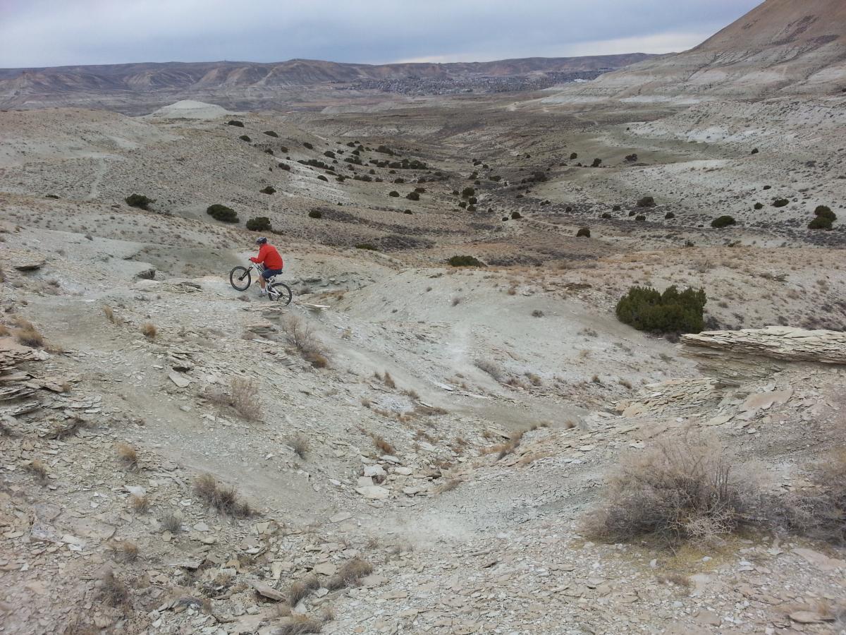 A mountain biker in a red jacket rides down a rocky slope in a barren landscape with distant hills and sparse vegetation. Tnt mountain bike trail.
