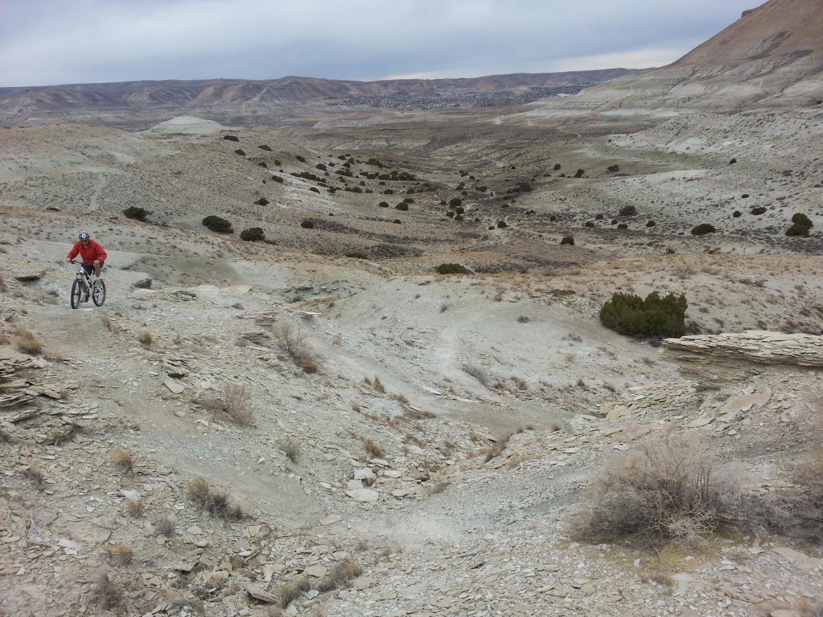 A mountain biker in a red jacket rides along a rocky trail in a barren landscape with rolling hills and sparse vegetation under an overcast sky. Tnt mountain bike trail.