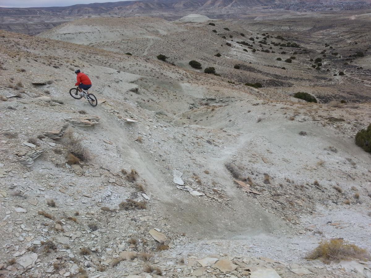 A person riding a mountain bike down a rocky, sloped trail in a rugged, barren landscape with rolling hills and sparse vegetation. Tnt mountain bike trail.