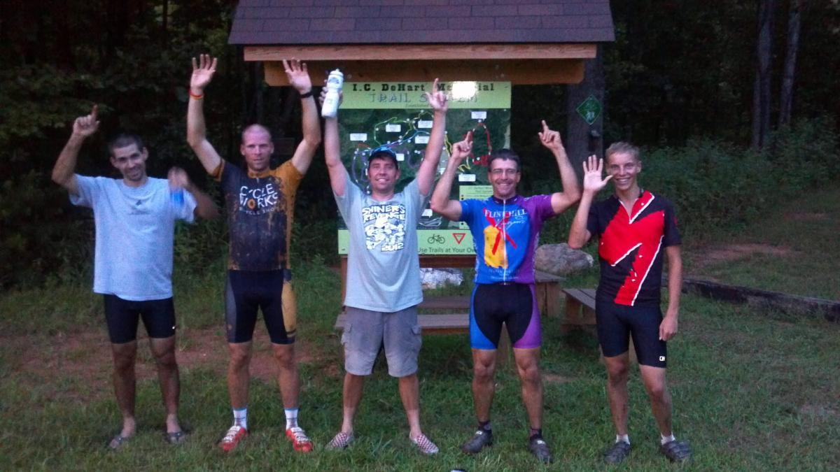 A group of five cyclists celebrating at a trailhead, raising their hands in a triumphant gesture. They are standing in front of a sign for I.C. DeHart Memorial Trail System, surrounded by greenery. The cyclists are wearing various cycling gear and appear to be in high spirits after a ride. Woolwine Trails [Shiners Revenge] mountain bike trail.