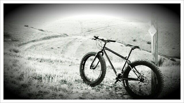 A black and white photo of a fat bike parked on a grassy hillside, with dirt trails winding in the background. A trail marker stands nearby, indicating outdoor recreational paths. The scene conveys a sense of adventure and the beauty of nature. Green Mountain mountain bike trail.