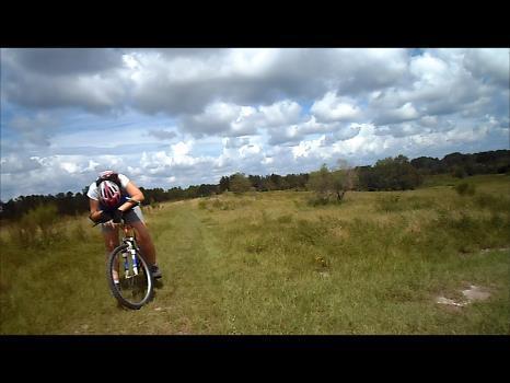 A cyclist riding a mountain bike on a grassy trail under a partly cloudy sky. The cyclist is leaning forward, focusing on the path ahead, while the surrounding landscape features open fields and patches of trees in the background. Balm Boyette Scrub Preserve mountain bike trail.