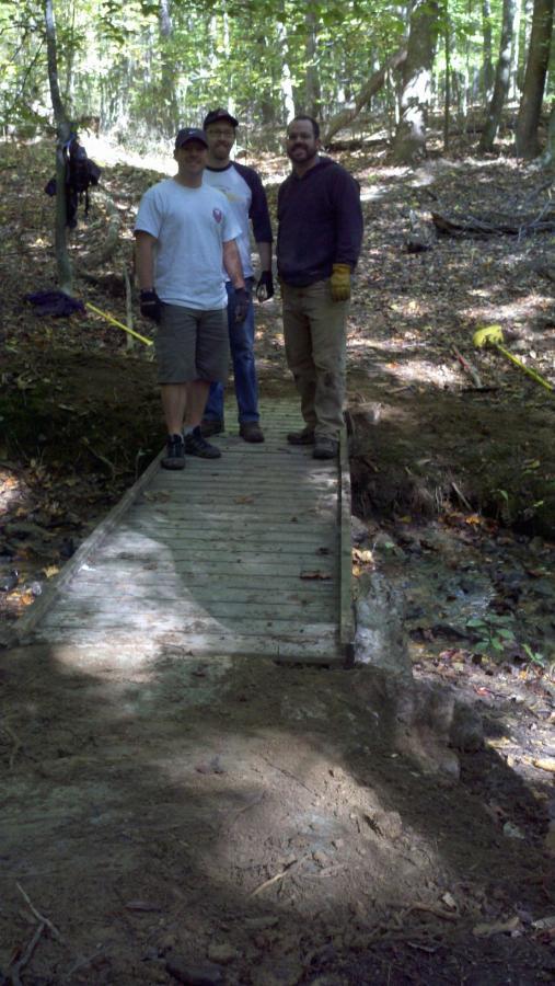 Three men stand on a wooden bridge in a forested area. The bridge crosses a small creek, and the ground around it is recently cleared of debris. They are wearing casual outdoor clothing, and the scene is illuminated by dappled sunlight filtering through the trees. In the background, a person can be seen working further down the trail. Freedom Center mountain bike trail.