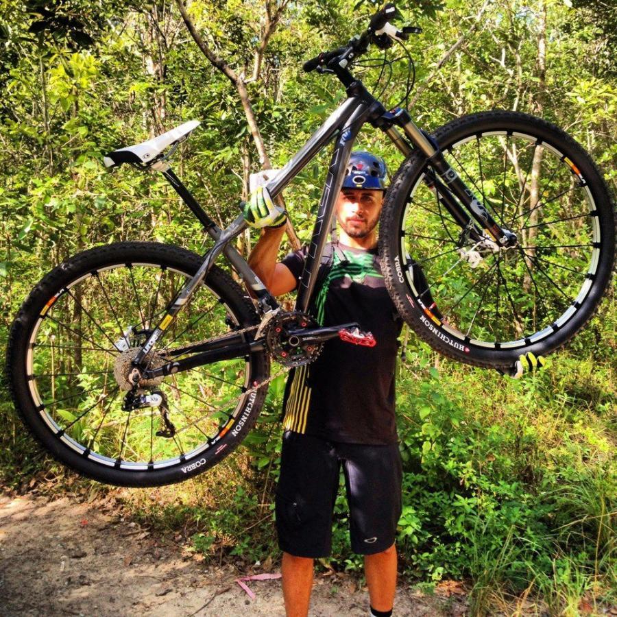 A cyclist in outdoor gear holds a mountain bike above his head, surrounded by greenery in a forested area. The cyclist wears a helmet, gloves, and a black shirt with green accents, while the bike features prominent wheels with detailed treads. Tortuguero mountain bike trail.