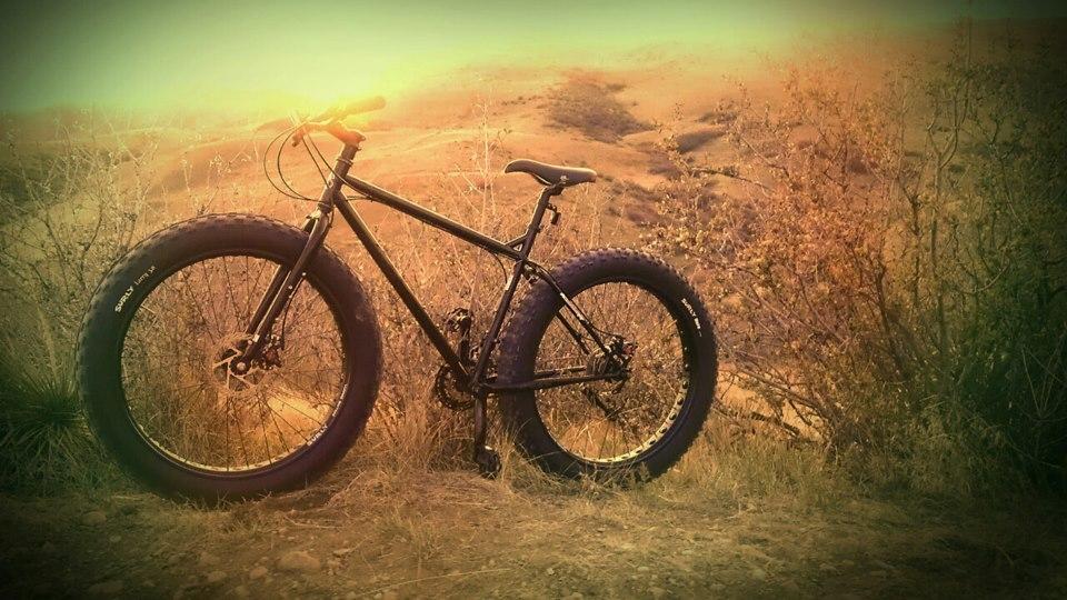A black fat bike resting on an uneven terrain with dry grass and sparse bushes in the background, under a hazy, sunlit sky. The bike features oversized tires and a rugged design, ideal for off-road cycling. Green Mountain mountain bike trail.