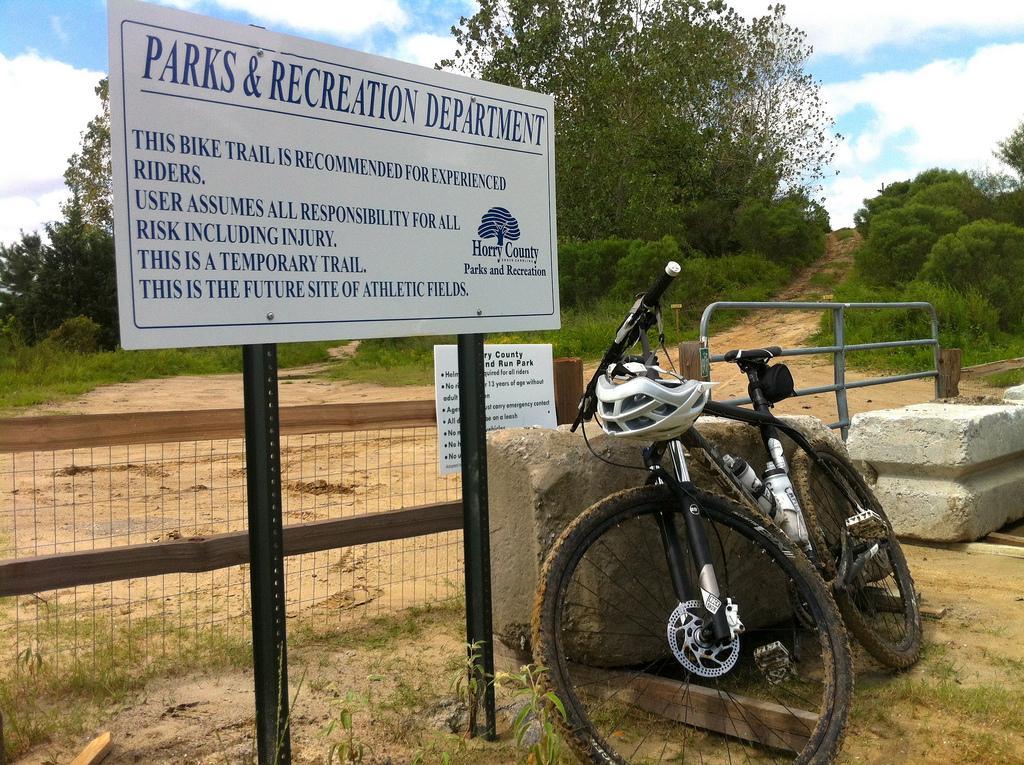 A sign for the Parks & Recreation Department indicating the rules for a bike trail recommended for experienced riders, accompanied by a mountain bike and helmet resting against a stone barrier. The background features a dirt path leading into a wooded area, with trees and vegetation visible. Horry County Bike Run Park mountain bike trail.