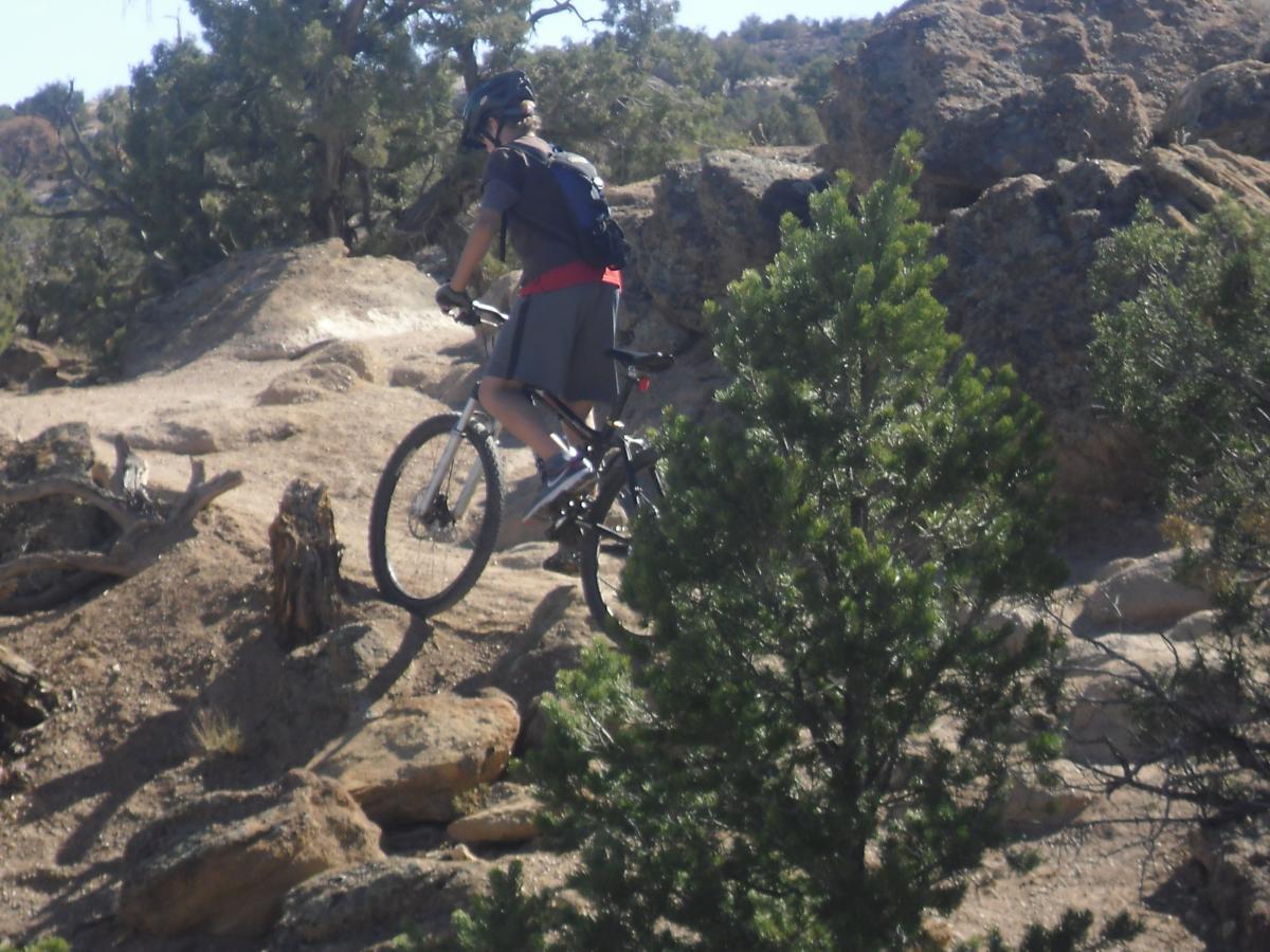 A person riding a mountain bike on a rocky trail surrounded by trees and boulders, in a natural outdoor setting. Pet - A - Kes mountain bike trail.