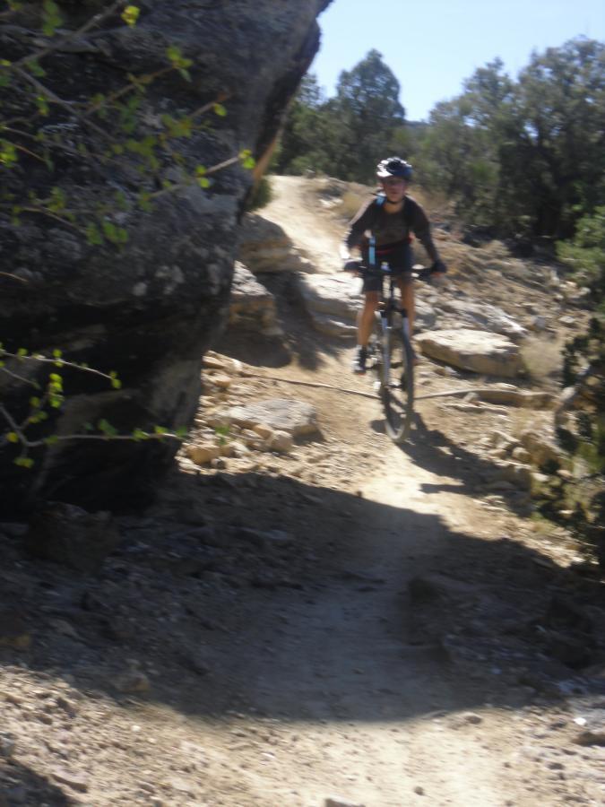 A mountain biker riding along a narrow dirt trail surrounded by rocky terrain and sparse vegetation on a sunny day. The biker is wearing a helmet and navigating around large boulders. Holy Cross mountain bike trail.