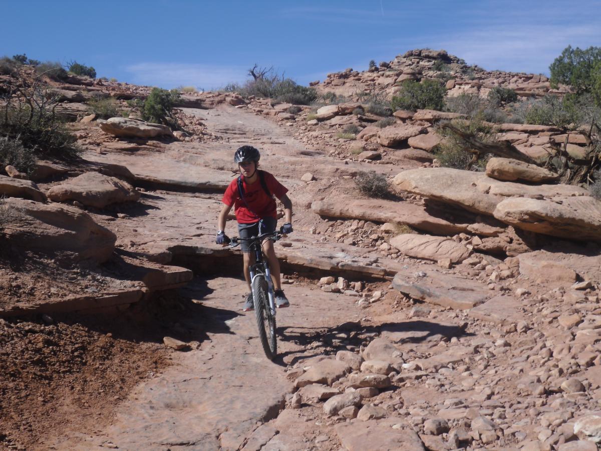 A young person cycling on a rocky trail in a desert landscape, surrounded by shrubs and rugged terrain under a clear blue sky. The cyclist is wearing a helmet and a red shirt while navigating the uneven path. Amasa Back Trail mountain bike trail.