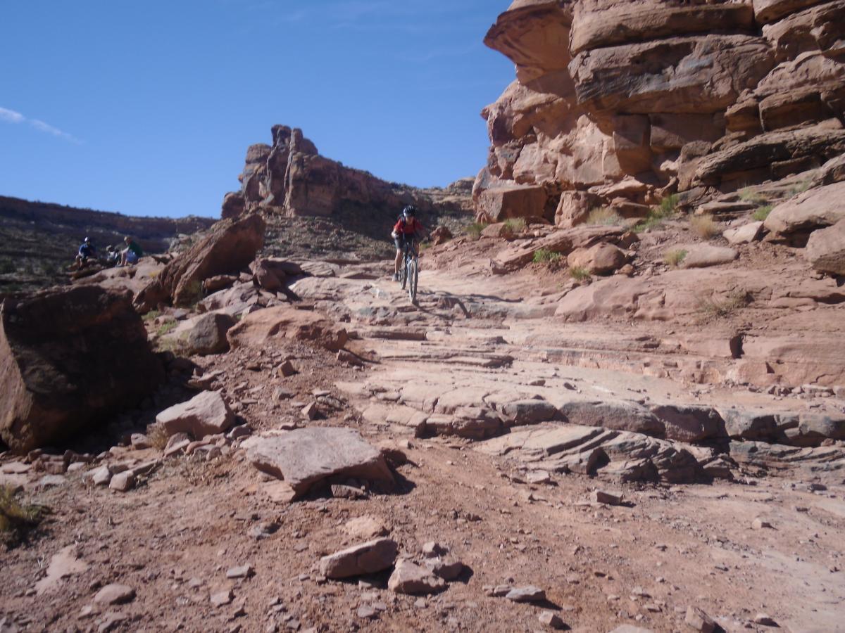 A mountain biker navigating a rocky trail surrounded by steep cliffs and boulders under a clear blue sky. Other bikers are visible in the background enjoying the rugged terrain. Amasa Back Trail mountain bike trail.