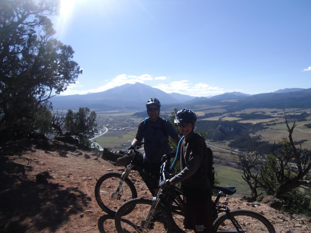Two mountain bikers pose for a photo on a scenic overlook, surrounded by natural landscapes. They're wearing helmets and biking gear, with bicycles beside them. In the background, rolling hills and distant mountains can be seen under a clear blue sky. Red Hill mountain bike trail.