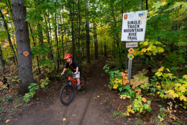 A young rider in a red helmet navigates a single track mountain bike trail surrounded by vibrant autumn foliage. The path features a sign indicating the trail and warning symbols nearby, with trees and colorful leaves creating a natural backdrop. Pinder mountain bike trail.