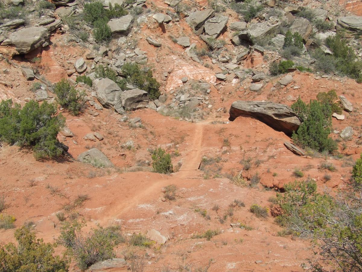 An arid landscape featuring reddish soil, scattered boulders, and patches of vegetation, with a winding dirt path leading through the terrain. Palo Duro Canyon mountain bike trail.