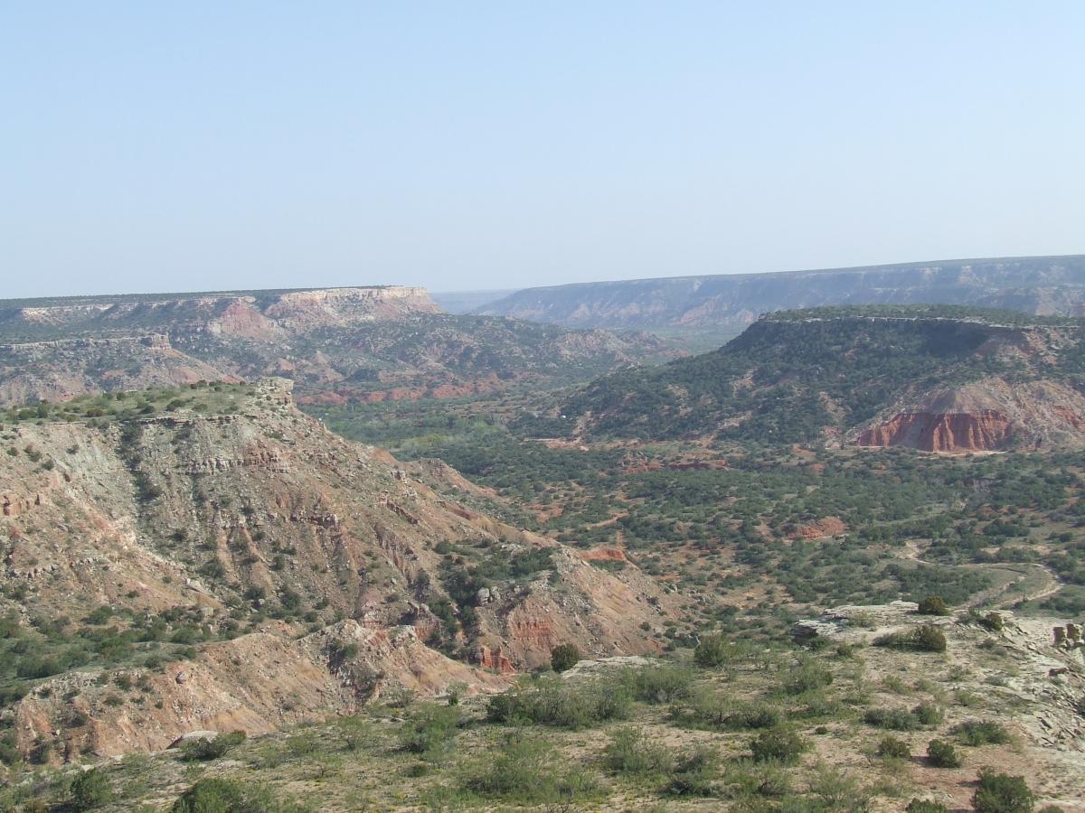 A panoramic view of a rugged canyon landscape featuring layered rock formations, sparse vegetation, and a clear blue sky. The terrain includes steep hills and valleys, showcasing earthy tones of red, brown, and green. Palo Duro Canyon mountain bike trail.