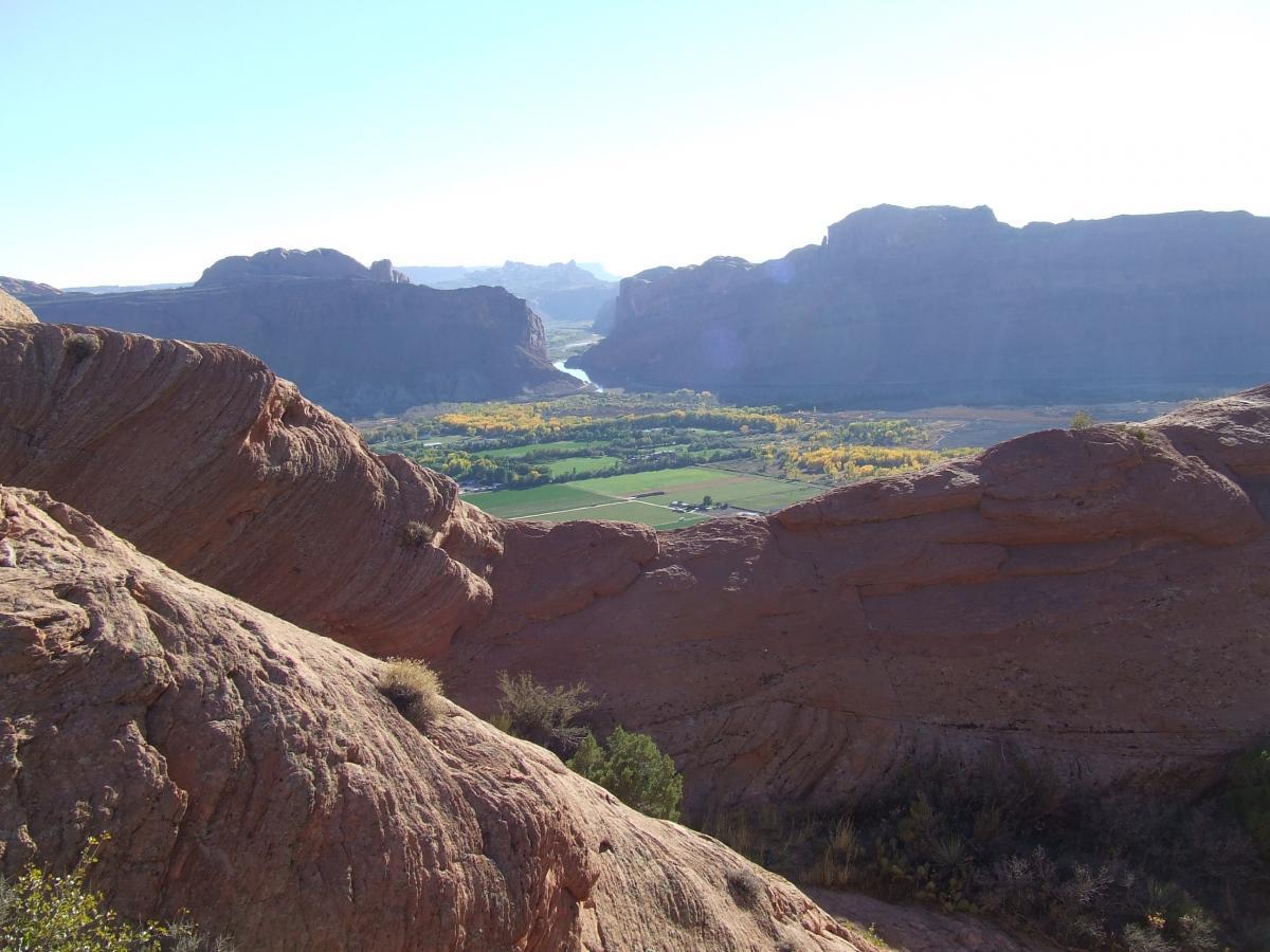 A panoramic view of a lush valley surrounded by rugged red rock formations and distant mountains, with a winding river and patches of green farmland below. The landscape is bathed in sunlight, highlighting the natural colors and textures of the rocks and vegetation. Slickrock mountain bike trail.