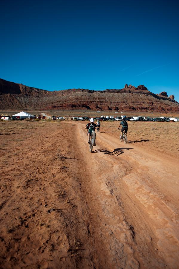 Two cyclists ride on a dirt path through a wide-open landscape, with a backdrop of rocky hills under a clear blue sky. In the distance, a campsite is visible, with tents and vehicles parked. The scene captures a sense of adventure and outdoor activity. Moab Brand Trails mountain bike trail.