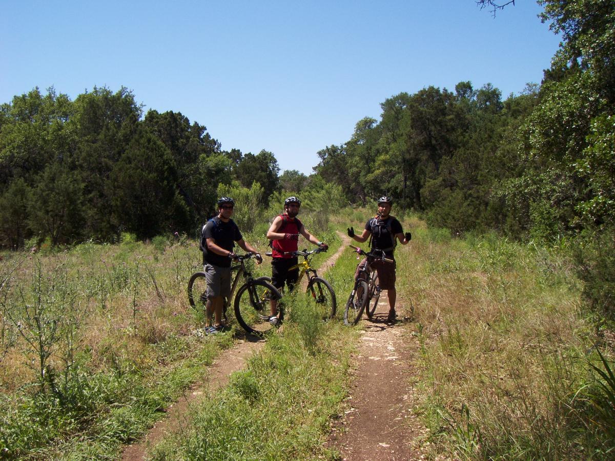 Three mountain bikers pause on a dirt path surrounded by tall grass and trees on a sunny day. They are smiling and wearing helmets, with two of them holding their bikes while the third gestures with his hands. The scene captures a moment of outdoor adventure and camaraderie. Salado Creek mountain bike trail.