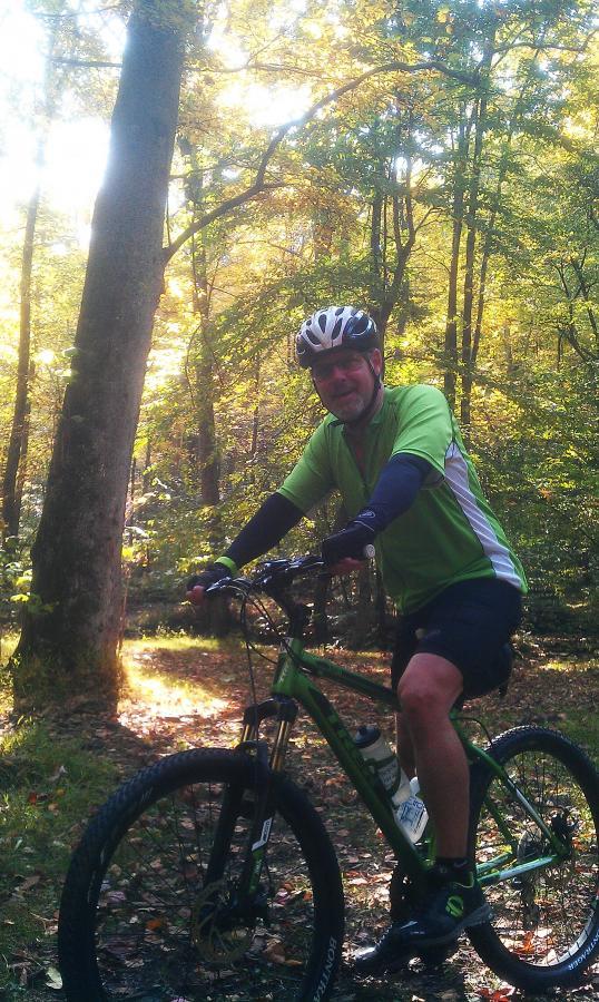 A cyclist in a green jersey and helmet poses on a mountain bike in a sunlit forest setting, surrounded by trees with autumn foliage. The scene captures the essence of outdoor recreation and adventure. Coopers Rock mountain bike trail.