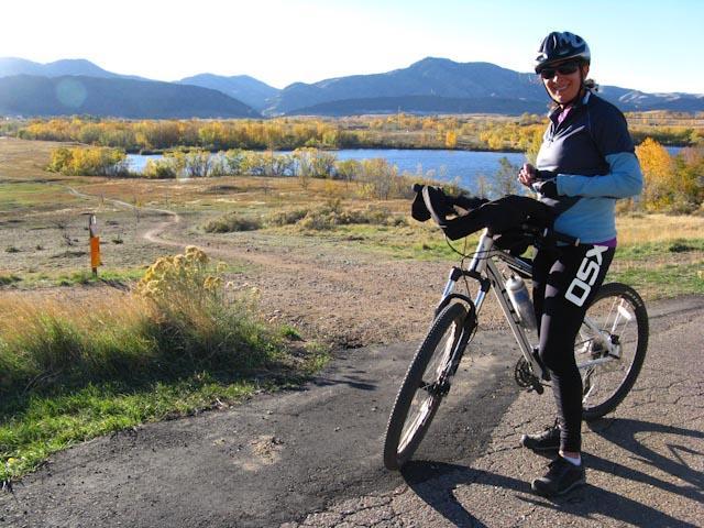 A woman wearing a helmet and cycling attire stands next to a mountain bike on a scenic trail. She is smiling, with a backdrop of colorful autumn foliage and a serene body of water, surrounded by mountains under a clear blue sky. Bear Creek Lake Park mountain bike trail.