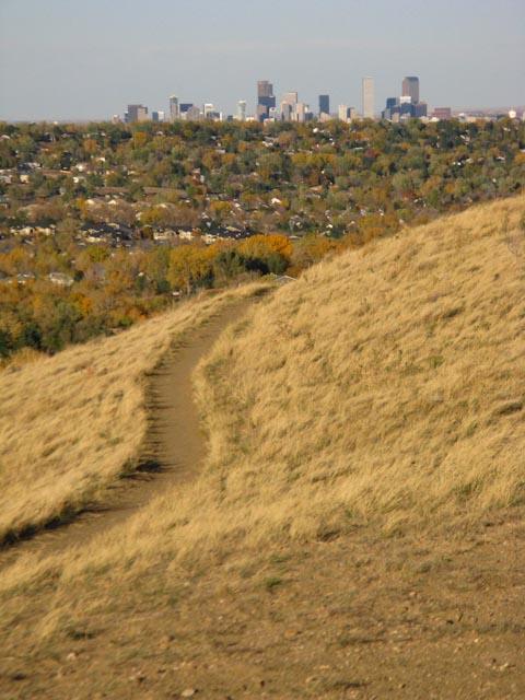 A winding dirt path leads through tall, golden grass on a hillside, with a panoramic view of a city skyline in the distance. The landscape features autumn foliage in varying shades of green and yellow, suggesting a mild season. Bear Creek Lake Park mountain bike trail.