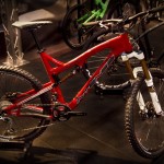 A modern mountain bike with a vibrant red frame, featuring a suspension fork, thick tires, and a sleek design. The bike is displayed in a showroom alongside other bicycles, set against a dark background.