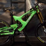 A vibrant green downhill mountain bike displayed on a stand, showcasing its sleek frame, specialized suspension components, and rugged tires designed for off-road terrain. The bike features a prominent brand logo and high-performance parts suitable for intense riding.