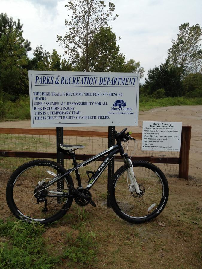 A mountain bike leaning against a wooden post in front of a sign for the Horry County Parks & Recreation Department. The sign details information about a bike trail recommended for experienced riders, including warnings about responsibility for risk and mentioning that the area will become a future site for athletic fields. Surrounding the area are trees and a dirt path. Horry County Bike Run Park mountain bike trail.