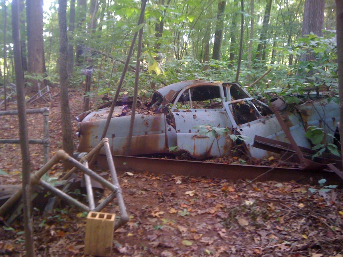 An abandoned, rusted car partially overgrown with vegetation in a wooded area, surrounded by trees and fallen leaves. Back Yard Trails mountain bike trail.