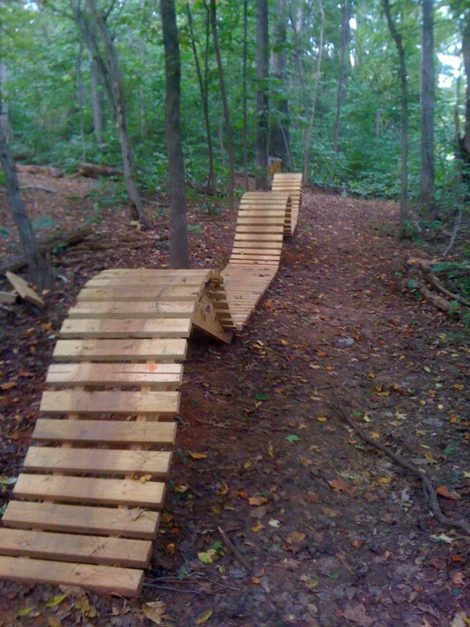 A wooden bike ramp curves along a dirt trail in a forested area, surrounded by trees and fallen leaves. The ramp features multiple sections with a slatted design, leading through the natural landscape. Back Yard Trails mountain bike trail.