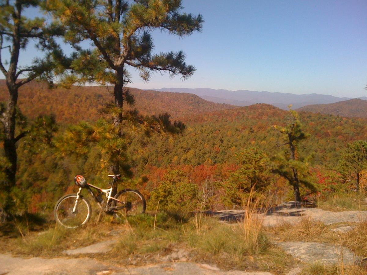 A scenic view of a mountainous landscape during autumn, featuring vibrant foliage in shades of red, orange, and green. In the foreground, a mountain bike is parked near a rocky outcrop, surrounded by pine trees. The clear blue sky adds to the idyllic outdoor setting. DuPont State Recreational Forest mountain bike trail.