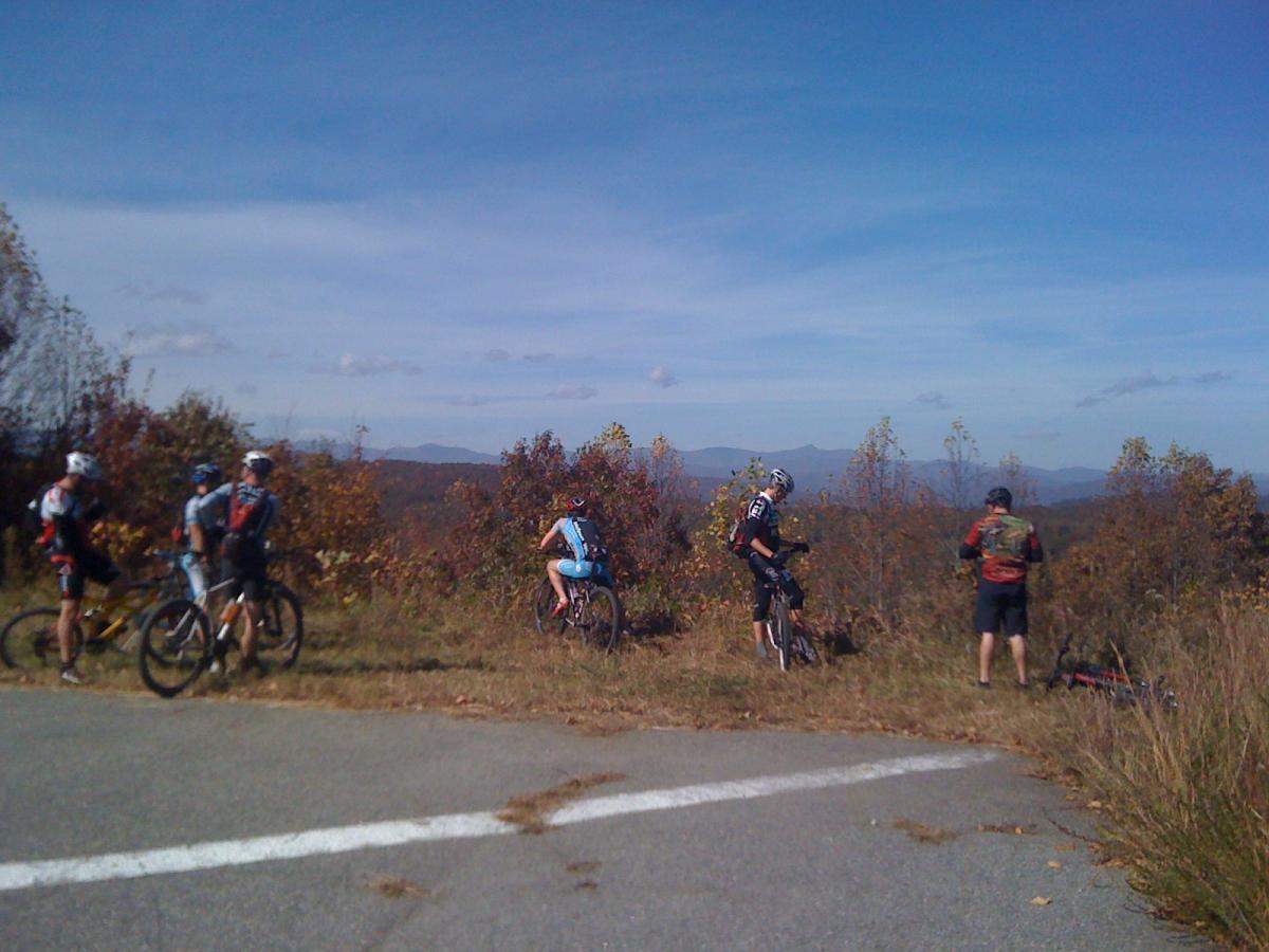 A group of five mountain bikers pause on a scenic overlook, surrounded by autumn foliage on a clear day. They are dressed in cycling gear, with some adjusting their bikes and others taking in the view of distant mountains under a blue sky. A winding road and patches of grass are in the foreground. DuPont State Forest mountain bike trail.