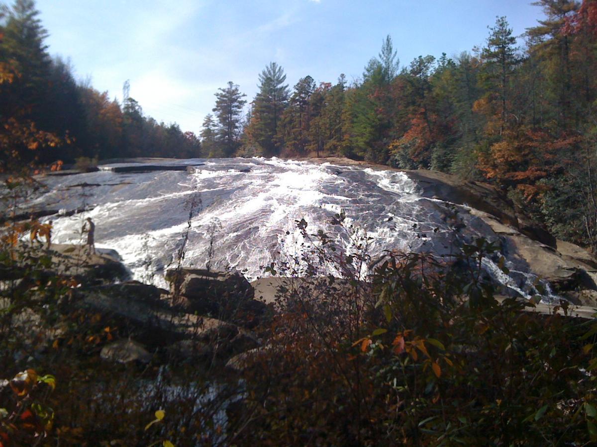 A serene view of a waterfall cascading down a smooth, rocky surface, surrounded by colorful autumn foliage and greenery. The sky is clear with soft clouds, creating a picturesque natural landscape. A person can be seen on the rocks near the water, enjoying the scenery. DuPont State Recreational Forest mountain bike trail.
