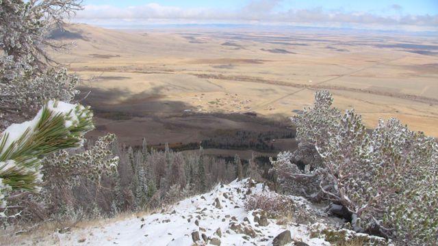 A scenic view of a snow-capped mountain slope leading down to a vast valley below, with patches of green and brown vegetation. The horizon features distant hills and a clear blue sky, partially filled with clouds. Snow lightly covers the ground in the foreground, emphasizing the elevation of the viewpoint. Centennial Ridge mountain bike trail.