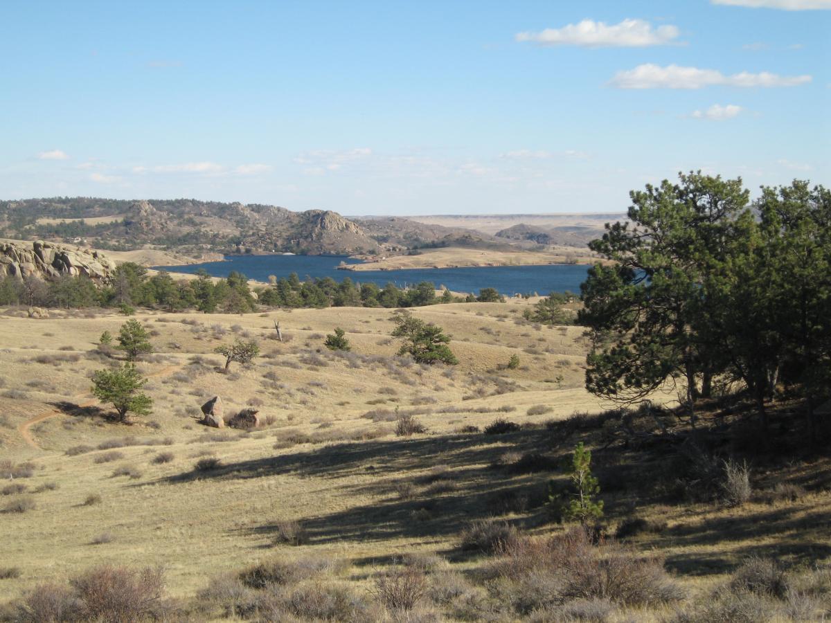A scenic landscape featuring a stony terrain with sparse vegetation, leading to a calm blue lake surrounded by rolling hills and distant mountains under a clear blue sky with a few clouds. Curt Gowdy State Park mountain bike trail.