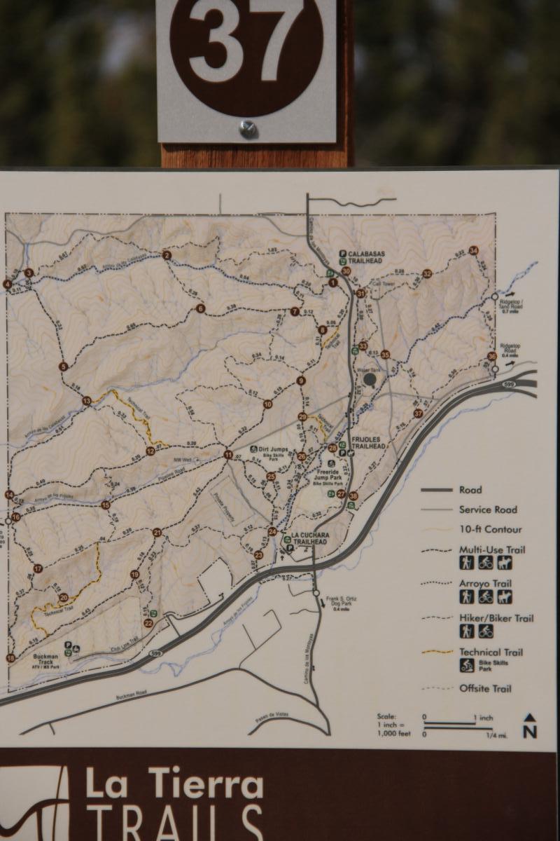 Map of La Tierra Trails, showing various trail routes including multi-use, hiker/biker, arroyo, and technical trails. The map is labeled with trail numbers and key points such as trailheads, with contour lines indicating elevation changes. A directional arrow marks north, and there is a scale for distance. La Tierra mountain bike trail.