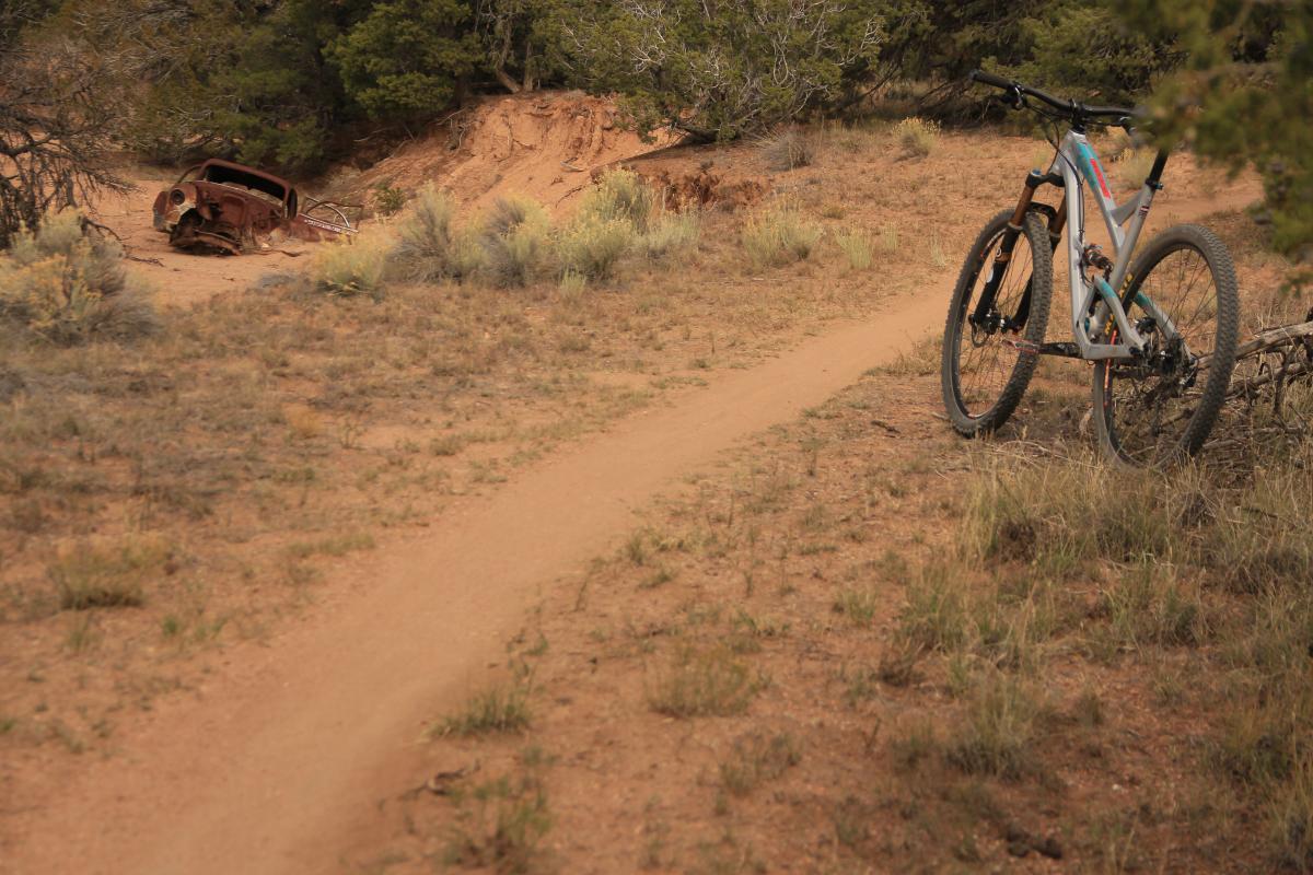 A mountain bike is resting beside a sandy dirt path in a rugged outdoor setting, with sparse vegetation and a weathered, abandoned car partially hidden in the background. La Tierra mountain bike trail.
