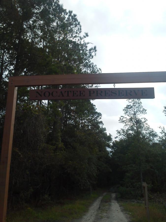 Wooden sign at the entrance of Nocatee Preserve, surrounded by trees, with a dirt path leading into the preserve. Nocatee mountain bike trail.