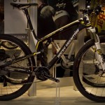 A sleek black and yellow mountain bike displayed on a booth, featuring a lightweight frame, disc brakes, and knobby tires designed for off-road performance. The backdrop includes promotional graphics related to cycling.