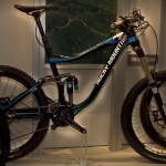 A blue and black mountain bike displayed in a showroom, featuring a sturdy frame, large tires, and advanced suspension components. The bike is positioned on a stand in front of a wall with a green landscape graphic.