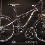 Mountain bike showcased on a display with a sleek design featuring a black and purple color scheme, high-quality suspension, and rugged tires. The frame prominently displays the "Pivot" brand logo. Background includes blurred promotional materials.