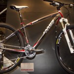 A black and red Jamis mountain bike displayed on a stand, featuring thick tires, a front suspension fork, and various gear components. The bike is showcased against a dark background, emphasizing its design and features.