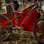 A high-performance mountain bike displayed in a natural setting, featuring a striking red and black frame, upgraded components, and fat tires. The bike is positioned on a wooden surface with foliage in the background, showcasing its rugged design ideal for outdoor adventures.