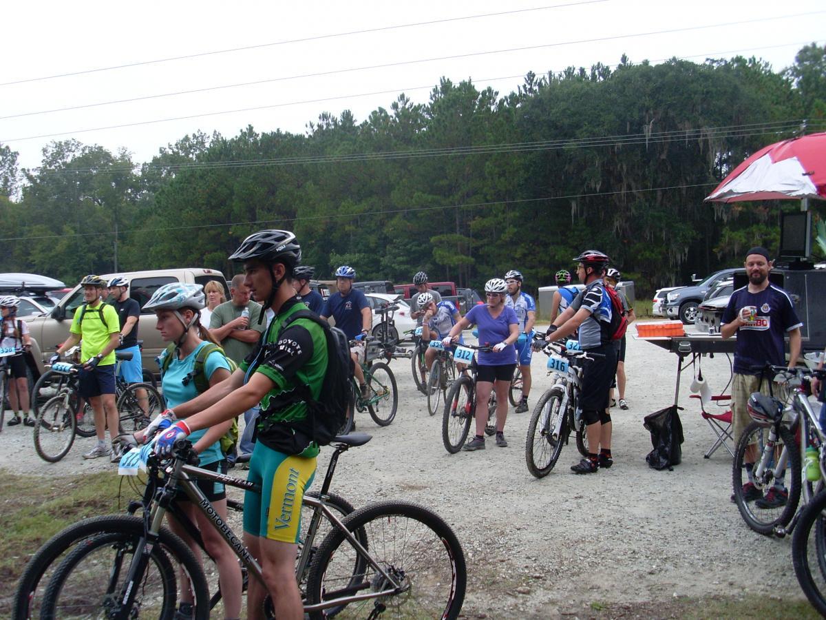 A group of mountain bikers gathers at the starting line of a race, wearing helmets and cycling gear. They are positioned near parked vehicles, with some spectators in the background. A food or refreshment station is visible to the right, under a small umbrella. The scene is set in a wooded area with trees in the background, suggesting an outdoor event. Wannamaker Park mountain bike trail.